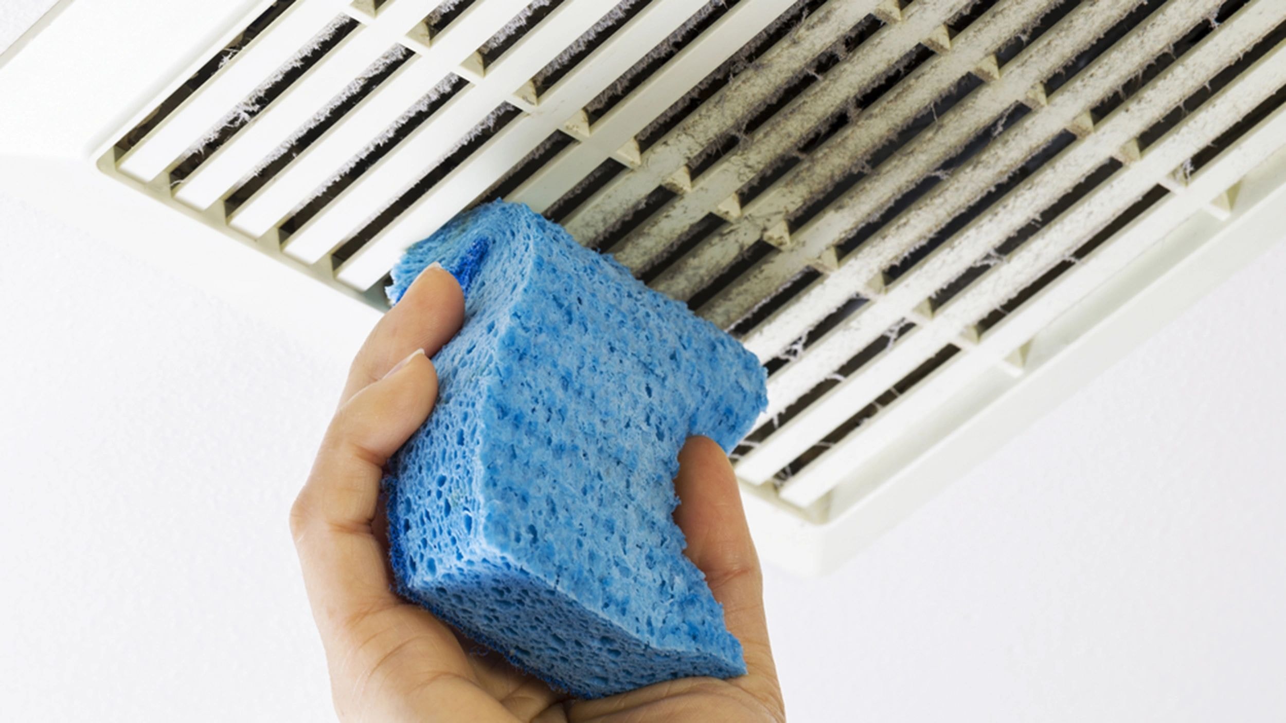 Hand holding a blue sponge near a dirty air vent — deep cleaning